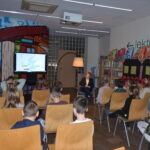 Grupa dzieci siedzi w sali, słuchając wykładu wyświetlanego na ekranie podczas zajęć bibliotecznych.