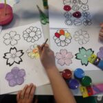 Dzieci malują farbami kontury kwiatów na papierze podczas warsztatów plastycznych.