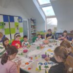 Dzieci siedzą przy stole i tworzą prace plastyczne z kolorowych elementów.