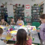 Dzieci uczestniczą w warsztatach plastycznych przy stoliku obłożonym materiałami i narzędziami do twórczej pracy.