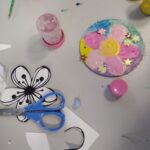 Na stole leżą wycięte z papieru kwiatki, nożyczki oraz przedmioty do rękodzieła, wykorzystane podczas warsztatów plastycznych dla dzieci.