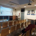 Sala szkoleniowa wyposażona jest w projektor, ekrany oraz szereg krzeseł ustawionych w rzędach.