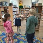 Trójka dzieci stoi na kolorowej macie w bibliotece otoczonej regałami z książkami.