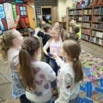 Grupa dzieci uczestniczy w zajęciach, stojąc wokół stołu pokrytego kolorowym materiałem.