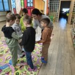 Grupa dzieci z dorosłą osobą uczestniczy w zajęciach na kolorowej macie, prawdopodobnie odgrywając scenkę związaną z handlem przyprawami.