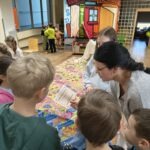 Grupa dzieci z nauczycielem ogląda kolorowe próbki przypraw rozłożone na stole podczas zajęć edukacyjnych.