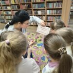 Grupa dzieci siedzi na dywanie w bibliotece, a kobieta pokazuje im dokument.