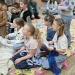 Grupa dzieci siedzi na kolorowej tkaninie, słuchając opowieści osoby stojącej z boku.