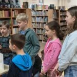 Grupa dzieci stoi w bibliotece, zainteresowana czymś widocznym poza klatką fotografii.