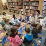 Grupa dzieci siedzi na dywanie w otoczeniu regałów z książkami podczas zajęć edukacyjnych.