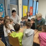 Grupa dzieci z nauczycielem obserwuje eksperyment z cieczą nienewtonowską podczas zajęć edukacyjnych.