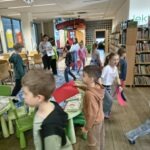 Grupa dzieci przemieszcza się po przestronnym pomieszczeniu z regałami pełnymi książek i elementami wystroju nawiązującymi do morskich podróży.