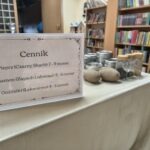 Na stole znajdują się przedmioty związane z handlem przyprawami oraz tabliczka z napisem "Cenik".