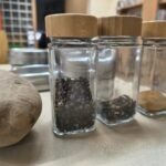 Trzy szklane słoiczki z drewnianymi nakrętkami wypełnione przyprawami stoją obok ziemniaka.