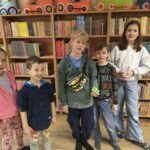 Grupa dzieci stoi przed regałami z książkami, prezentując elementy kostiumów nawiązujące do epoki odkryć geograficznych.
