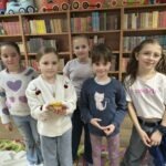 Pięć dziewczynek trzyma w dłoniach przedmioty związane z tematem zajęć, stojąc przed regałami pełnymi książek.