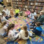 Grupa dzieci siedzi na kolorowej macie, otoczona regałami z książkami, podczas zajęć edukacyjnych.