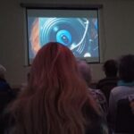 Na ekranie projekcyjnym wyświetlana jest abstrakcyjna kompozycja wizualna z dominującymi odcieniami błękitu i pomarańczy.