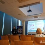 W sali konferencyjnej prezentowane są informacje o bezpieczeństwie cyfrowym na dużym ekranie, a uczestnicy siedzą w żółtych krzesłach.