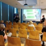 W sali konferencyjnej uczestnicy słuchają prezentacji na temat bezpieczeństwa cyfrowego, wyświetlanej na ekranie za prelegentami.