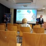 Na ekranie projekcyjnym widoczna prezentacja dotycząca bezpieczeństwa cyfrowego, a sala wypełniona jest uczestnikami spotkania.