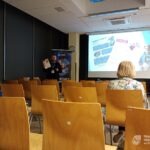 Mężczyzna prezentuje slajd podczas szkolenia dla seniorów, a za nim widoczne są puste krzesła i ekran projekcyjny.