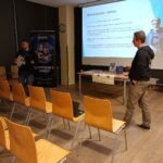 Mężczyzna prezentuje wykład przed publicznością siedzącą w sali konferencyjnej, a na ekranie wyświetlane są slajdy dotyczące bezpieczeństwa cyfrowego.