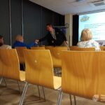 W sali konferencyjnej uczestnicy słuchają prezentacji eksperta, a na ekranie widoczna jest prezentacja multimedialna.