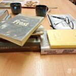 Na stole leżą sterty książek, notatnik oraz kubki z kawą.