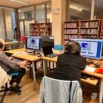 Uczestnicy warsztatów cyfrowych pracują przy komputerach w bibliotece, korzystając ze słuchawek.