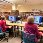 Uczestnicy warsztatów cyfrowych pracują przy komputerach ustawionych w bibliotece, w tle widoczne regały z książkami.