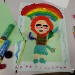 Dziecięca praca przedstawia postać z rudymi włosami stojącą pod tęczowym łukiem na zielonym tle.