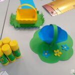 Wyklejanki przedstawiają papierowe koszyczki z rączkami umieszczone na zielonych podkładkach imitujących łąkę, ozdobione kwiatami.