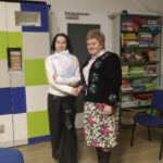 Dwie kobiety uściskiwają dłonie na tle regału z książkami i kolorowej dekoracji ściennej.