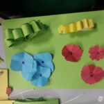 Na zielonym tle znajdują się papierowe dekoracje w kształcie kwiatów, koszyczków i uszu słonia.