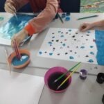 Dzieci malują farbami na papierze podczas warsztatów plastycznych w bibliotece.