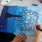 Dziecko rysuje czarny pień drzewa z białymi okrągłymi liśćmi na niebiesko-granatowym tle.