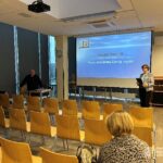 Kobieta stoi przy ekranie, prezentując slajd podczas wykładu w sali konferencyjnej.