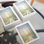 Na stole leżą trzy reprodukcje obrazów przedstawiających ścieżkę wijącą się wśród drzew.