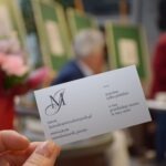 Ręka trzyma wizytówkę z logo i danymi kontaktowymi firmy, w tle widoczne osoby i zielone płachty.