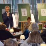 Mężczyzna w garniturze przemawia do publiczności, a za nim widoczne są dwa duże panele z grafikami.