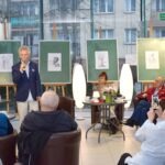 Mężczyzna w marynarce przemawia do publiczności, a kobieta siedzi przy stole otoczonym lampami.