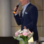 Mężczyzna w garniturze trzyma mikrofon i stoi obok bukietu kwiatów.