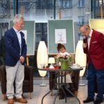 Dwóch mężczyzn rozmawia w pobliżu dziecka siedzącego przy stoliku z lampką i kwiatami podczas spotkania Akademii Słowa.