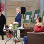 Mężczyzna w garniturze stoi tyłem, a przed nim stół z kwiatami i filiżankami, obok stoją dwa sztalugi z obrazami.