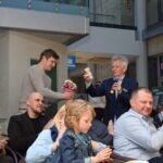 Mężczyzna w garniturze przemawia do publiczności, a obok niego stoi osoba podająca mu książkę.