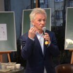 Mężczyzna w marynarce trzyma mikrofon i przemawia do publiczności, stojąc przed dwoma sztalugami z obrazami.