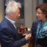 Mężczyzna w garniturze rozmawia z kobietą noszącą okulary, a w tle widoczna jest postać dziecka.