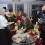 Kobieta w zielonym swetrze przegląda książki leżące na stoliku kawowym podczas spotkania Akademii Słowa.