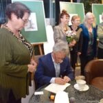 Mężczyzna siedzący przy stole podpisuje dokument, a w tle stoją kobiety obserwujące obrazy na sztalugach.